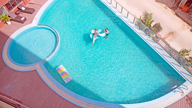 An aerial view of a swimming pool with two people on a float, surrounded by a deck with lounge chairs and potted plants. - City Comfort Airport Hotel Phnom Penh (Phnom Penh)