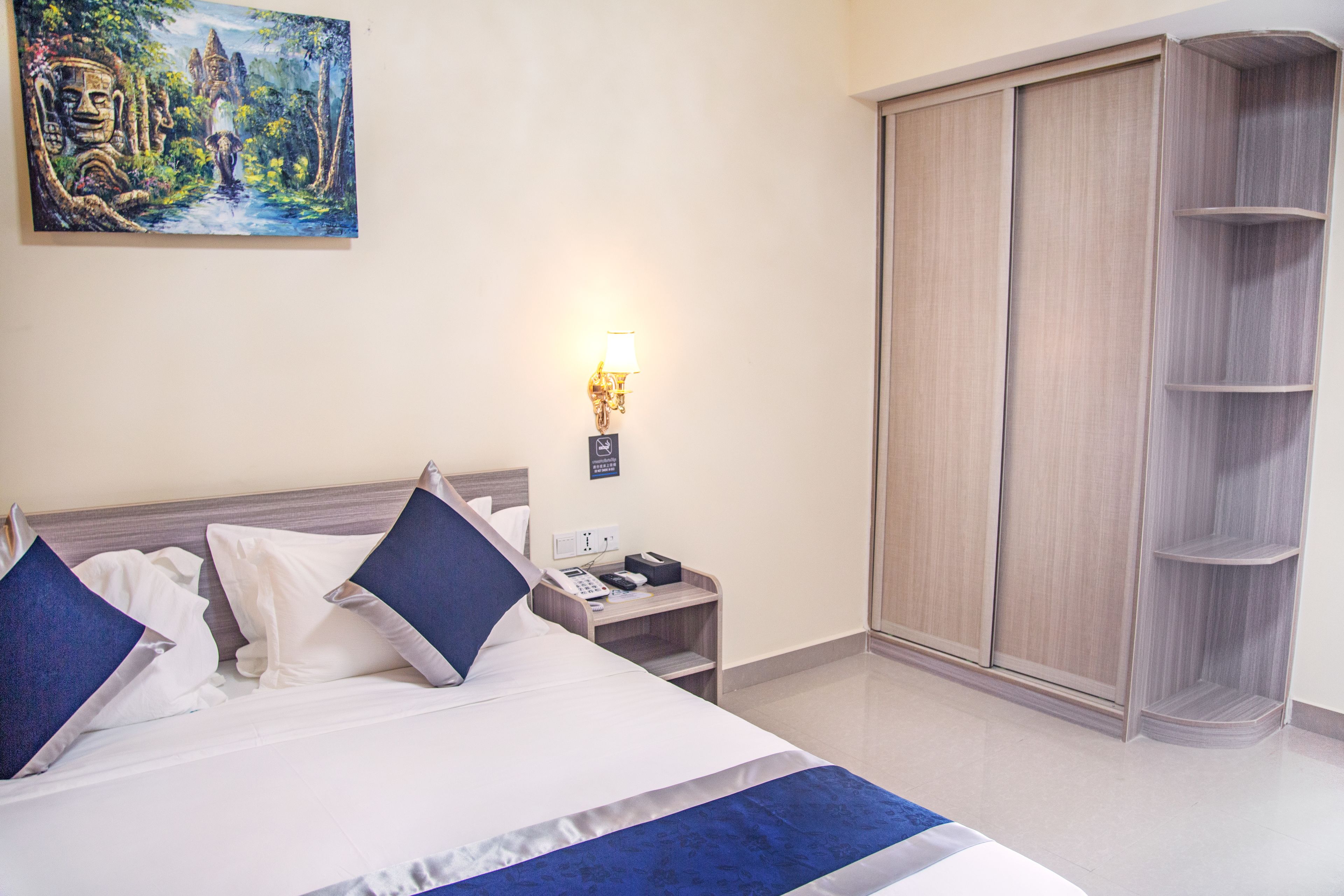 A hotel room with a bed, nightstand, lamp, wardrobe, and a painting on the wall.
