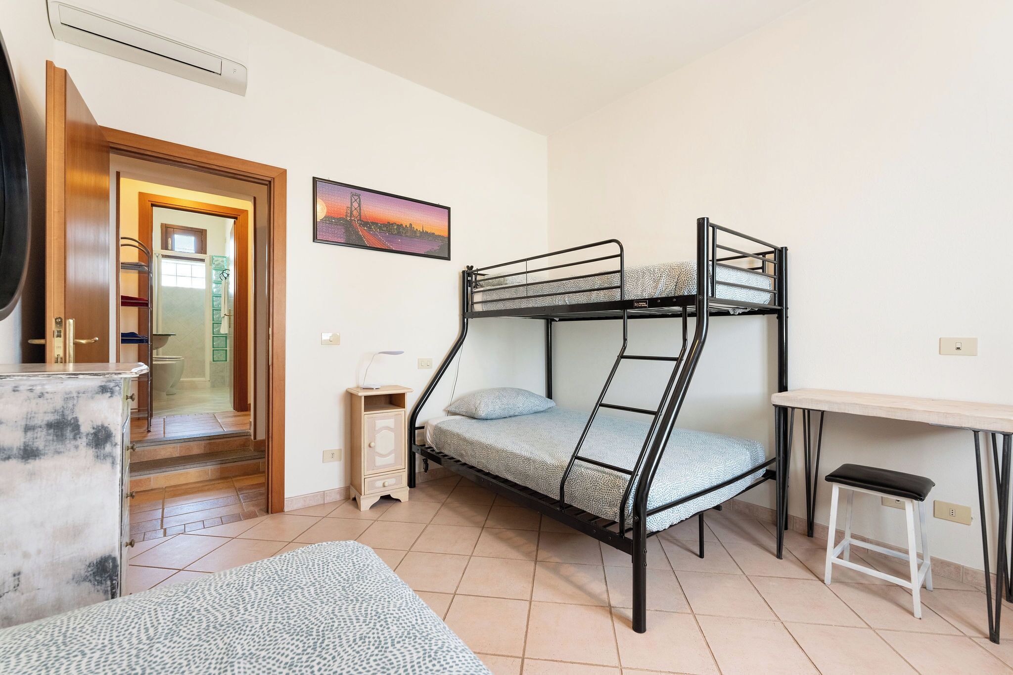 A bunk bed with a desk and a stool, a small cabinet, and a painting on the wall.