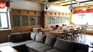 A living area with a sectional sofa, wooden dining table, and chairs. There are windows with curtains, a radiator, and a ceiling with wooden beams and chandeliers. - Gruppenhaus Rügen Zur Brunnenaue (Sagard)