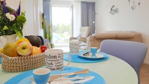 A dining area with a table set for two, featuring a basket of fruits, a mug with text, and a vase of flowers.