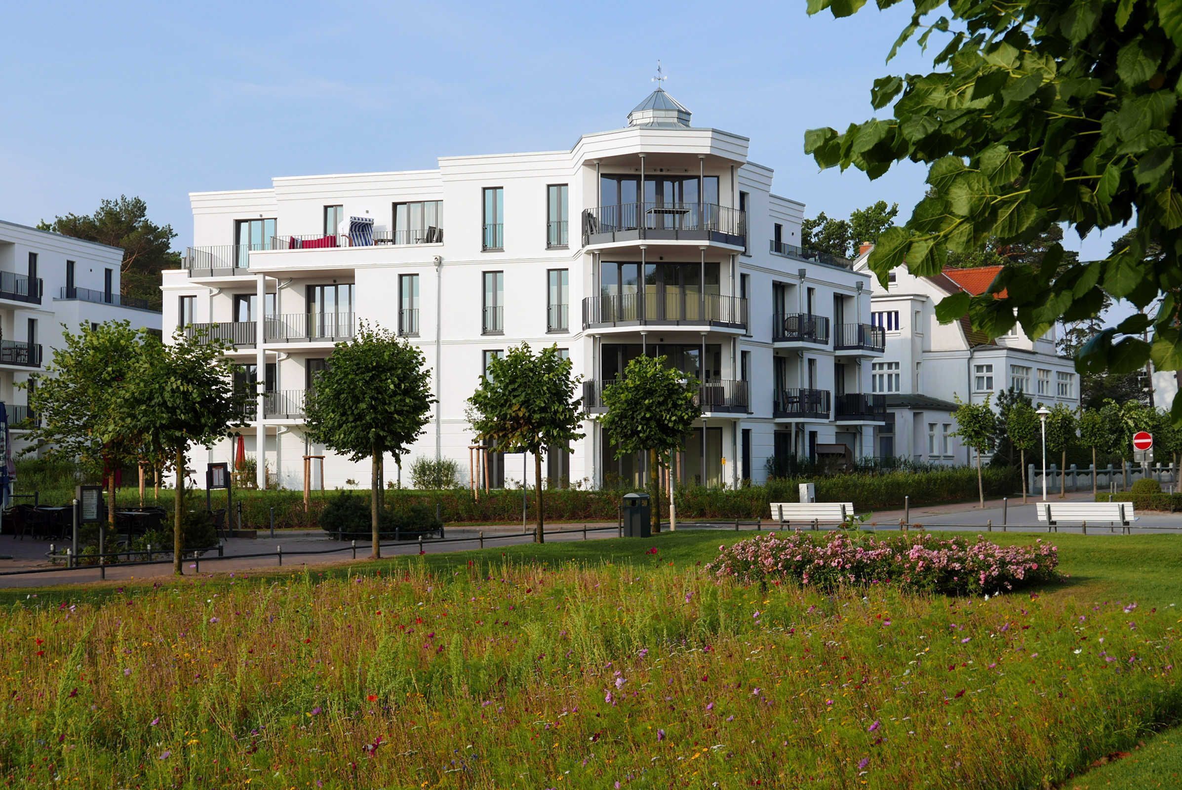A modern multi-story building with balconies, a landscaped garden, and a clear sky.