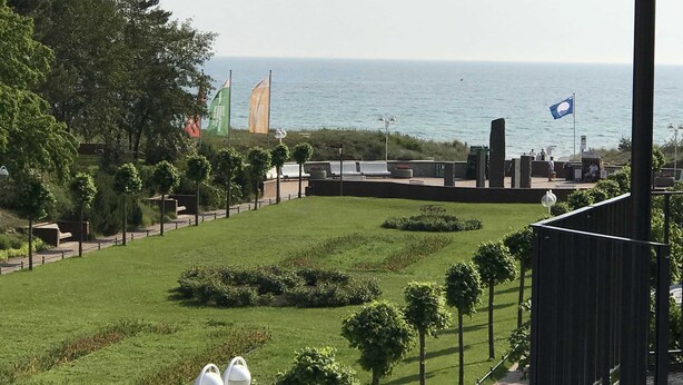 A landscaped area with a view of the sea, featuring a grassy lawn, trees, and a seating area with a flag.