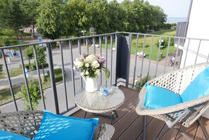 A balcony with wicker furniture, a small table with a vase of flowers, and a view of a park and street.