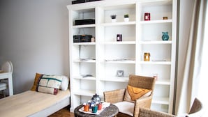 Una habitación pequeña y ordenada con una cama individual, una silla de mimbre, una mesita con una bandeja de objetos y un estante blanco adosado a la pared.