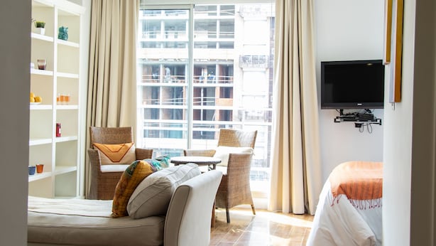 Una habitación de hotel moderna con sofá, sillón, televisor, estantería y un amplio ventanal con cortinas.