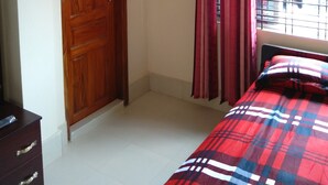 Une chambre avec une armoire en bois, un lit recouvert d’une couverture à carreaux, une fenêtre avec des barreaux en métal et un climatiseur fixé au mur.