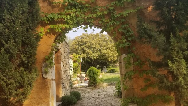 An arched stone pathway leading through a rustic garden with greenery and a clear sky. - Le Mas des Escans (Le Beausset)