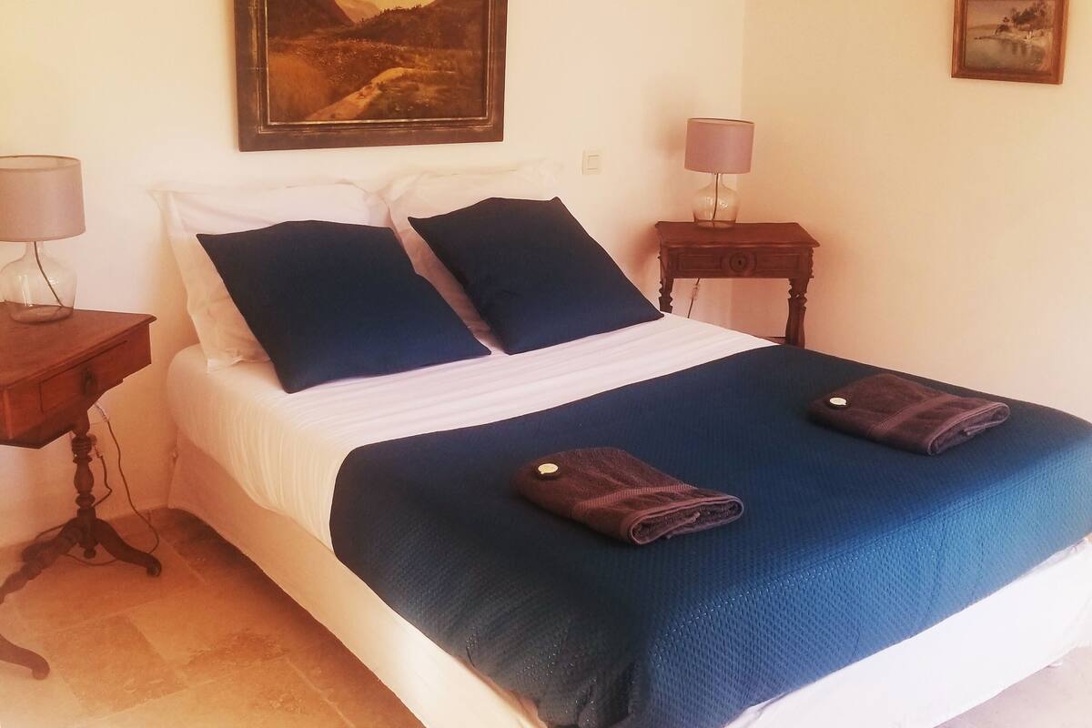 A neatly made bed with a blue cover, two pillows, and a folded towel on it. There are two bedside tables, each with a lamp. A painting is on the wall above the bed.