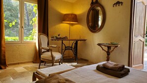 A room with a bed, a chair, a table with a lamp, a mirror, and a view of greenery outside. - Le Mas des Escans (Le Beausset)