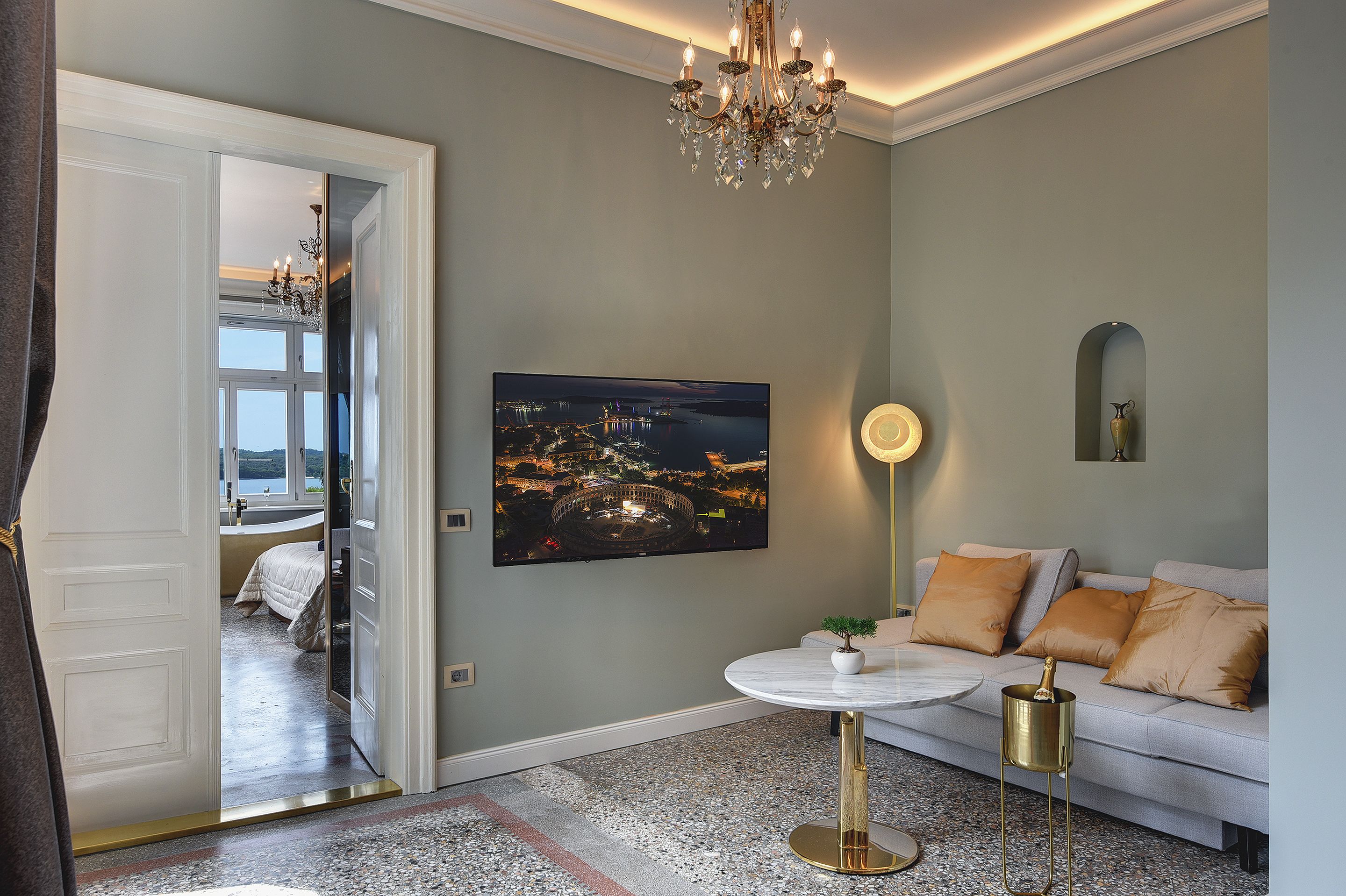 A modern living room with a large flat-screen TV, a round coffee table, a sofa with cushions, and a chandelier.