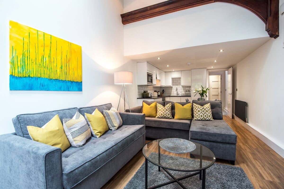 A modern living room with grey sofas, yellow and patterned cushions, a glass coffee table, and a large abstract painting on the wall.