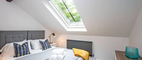 A bedroom with a skylight, a bed with towels, a bedside table with a lamp, a radiator, and a wooden dresser.