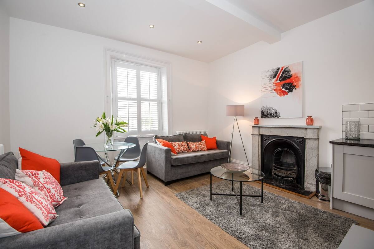 A modern living room with a grey sofa, red and white cushions, a glass dining table, and a fireplace.