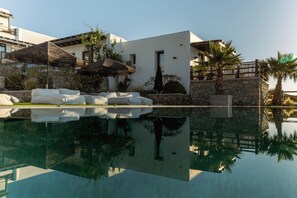 A pool with a clear reflection, a white building with a stone wall, and palm trees. - Nami by Maregold Mykonos, Adults Only (Mykonos)