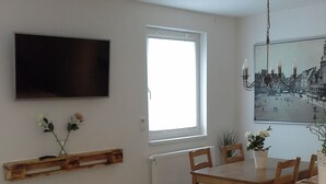 Un comedor con mesa y sillas de madera, televisor montado, ventana con persianas y un radiador.