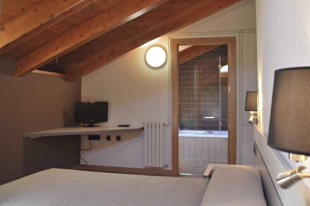 A bedroom with a wooden ceiling, a bed, a TV, and a window with blinds.