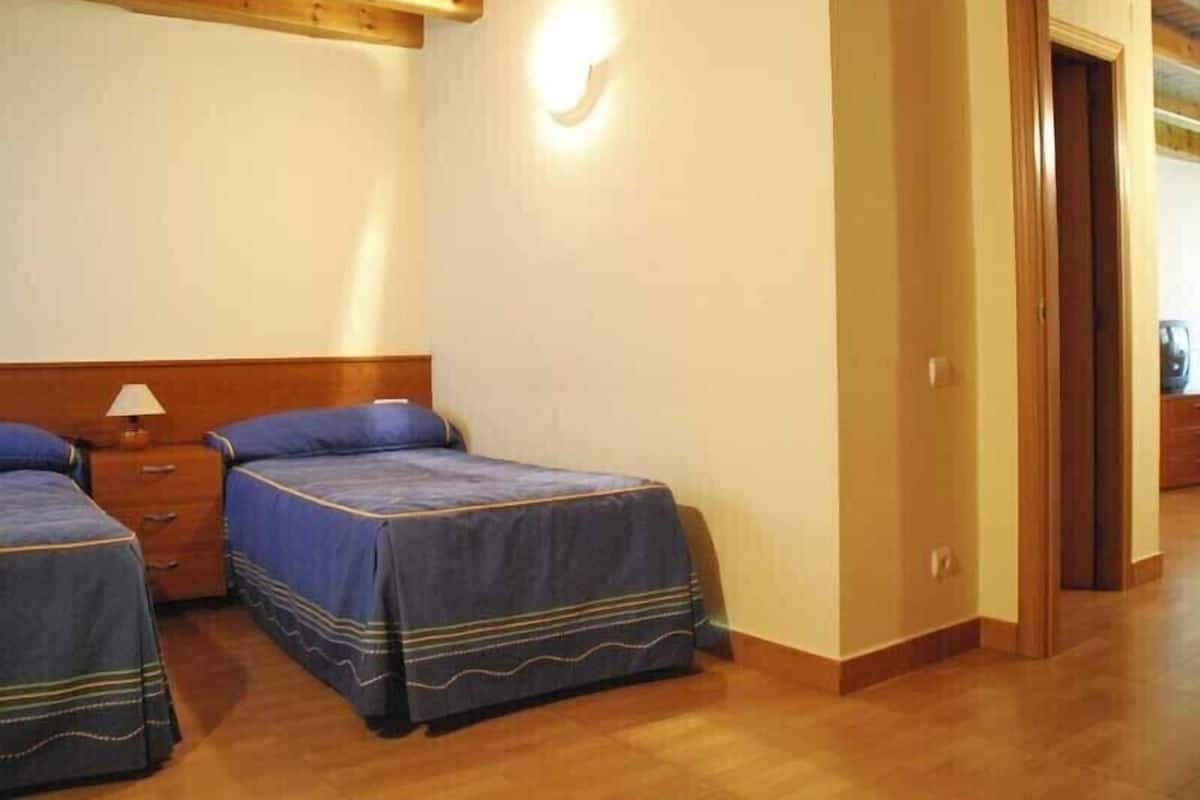 A hotel room with two beds, wooden ceiling beams, a nightstand with a lamp, and a door leading to another room.