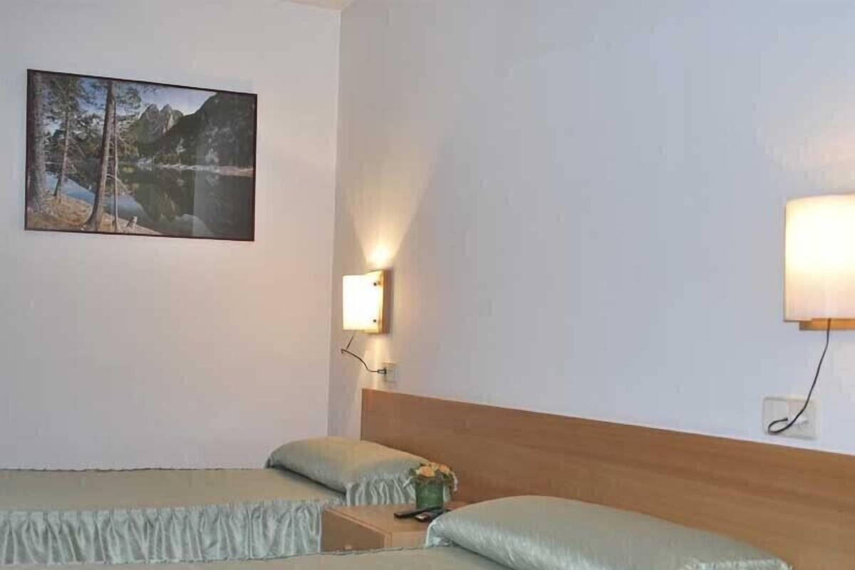 A hotel room with two beds, a painting of a mountain landscape, and a wall-mounted light fixture.