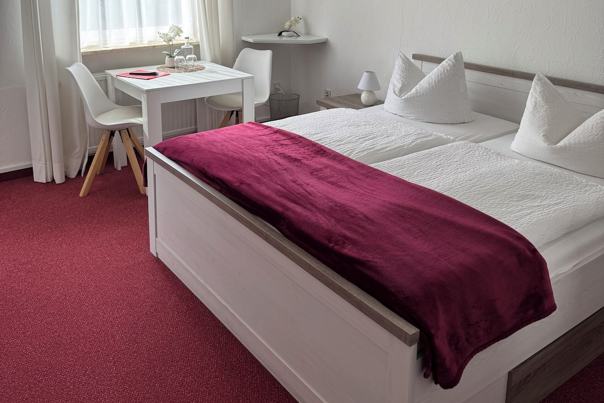 A hotel room with a bed, a desk, a chair, a window with curtains, and a painting on the wall.