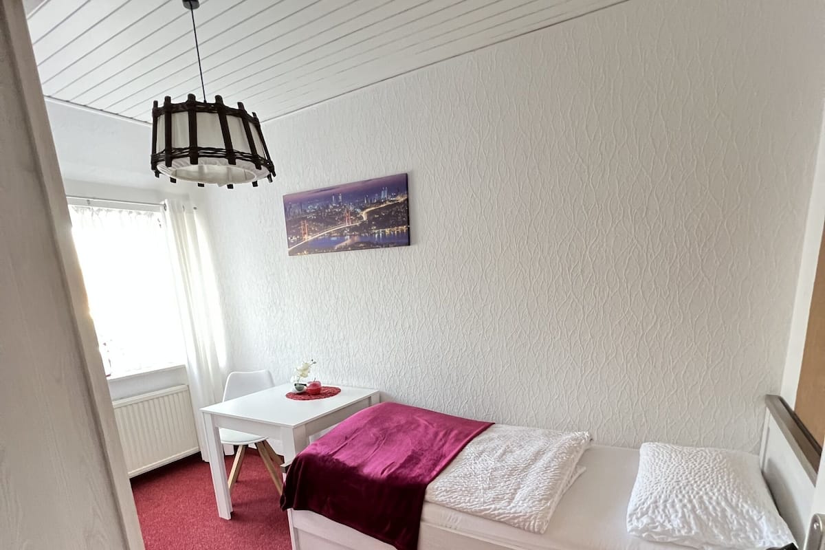 A bedroom with a bed, a desk, and a chair. There is a window with curtains, a hanging light fixture, and a painting on the wall.