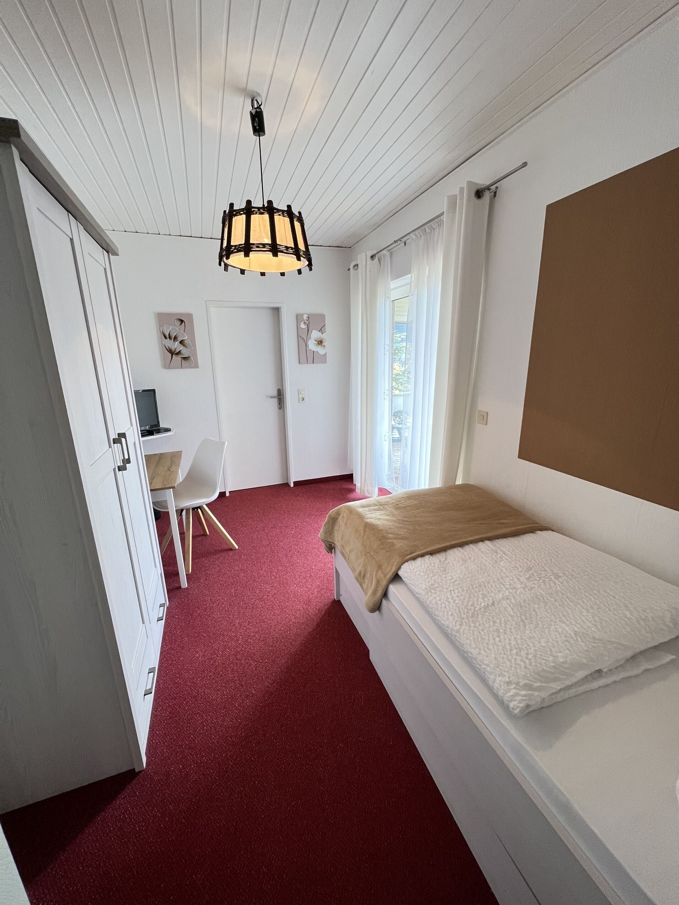 A bedroom with a single bed, a desk, and a chair. There is a door on the left and a window on the right.