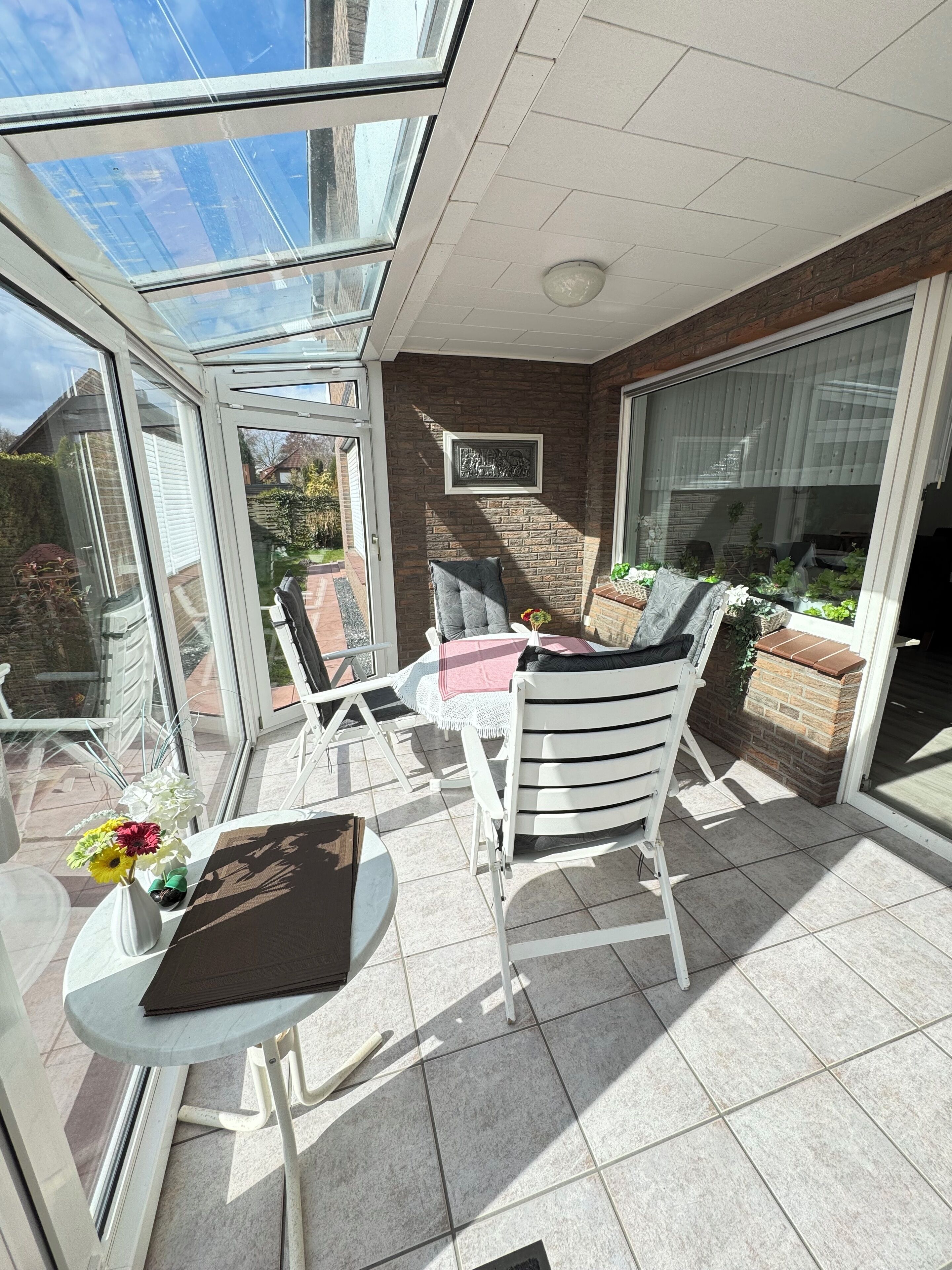 A sunlit patio with a glass door, a table with a vase of flowers, and a chair with a cushion.