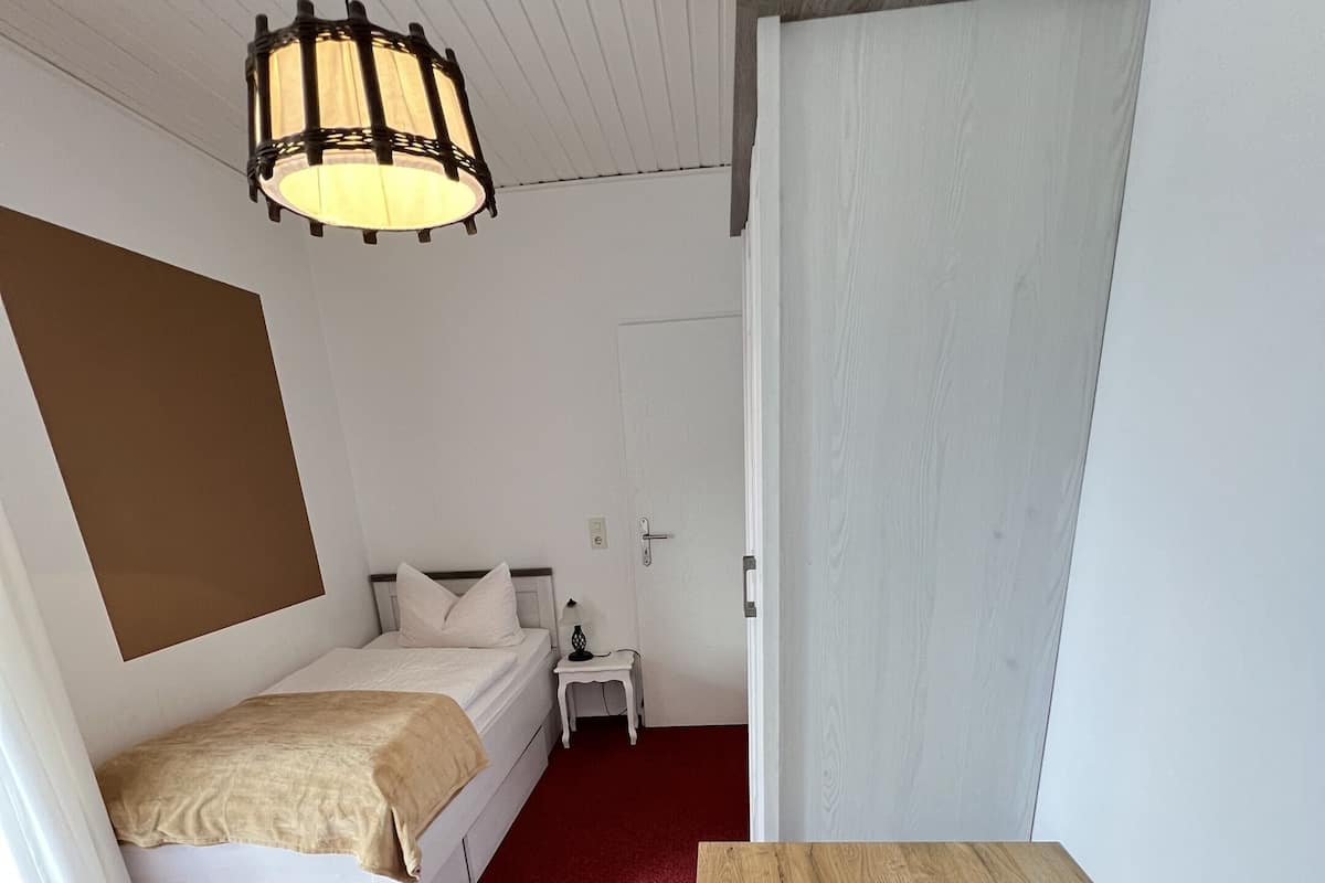 A compact room with a bed, a small table, a chair, and a hanging light fixture.