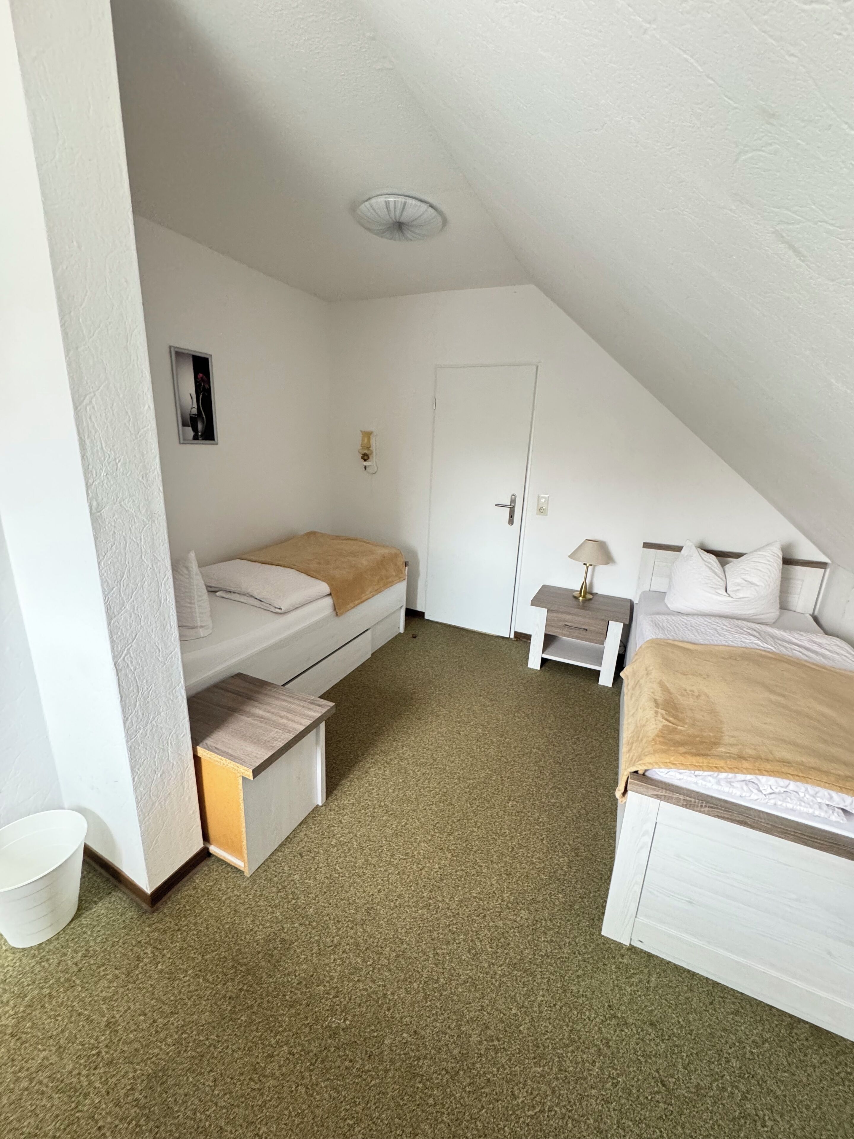Un dormitorio pequeño de estilo ático, con paredes blancas, techo inclinado, una cama individual, una mesita de noche y un mueble pequeño.