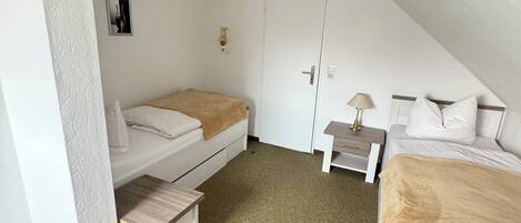 Un dormitorio pequeño de estilo ático, con paredes blancas, techo inclinado, una cama individual, una mesita de noche y un mueble pequeño.