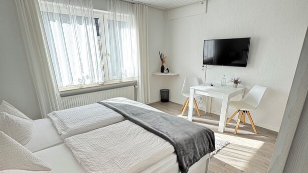 A modern bedroom with a bed, a TV, a small desk, and a window with curtains. - Hotel Pension Petra (Bad Zwischenahn)