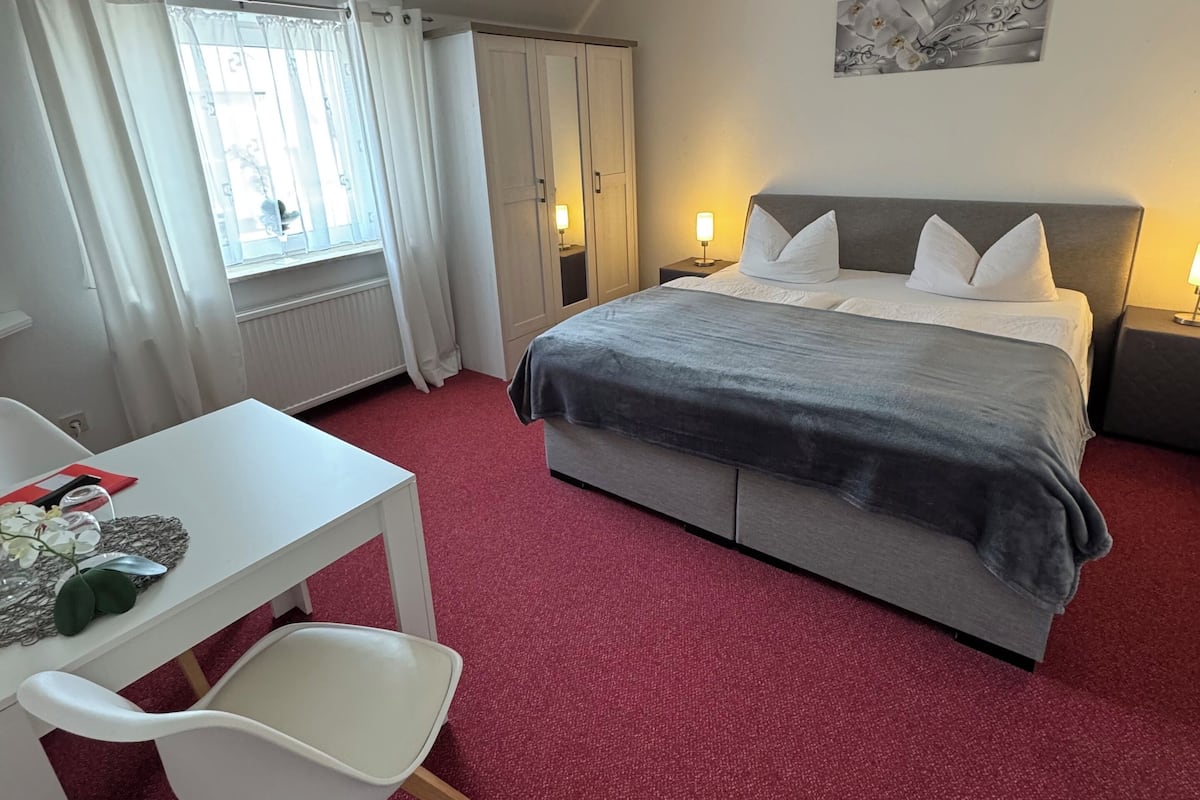 A hotel room with a bed, a desk, a chair, a window with curtains, and a ceiling light.