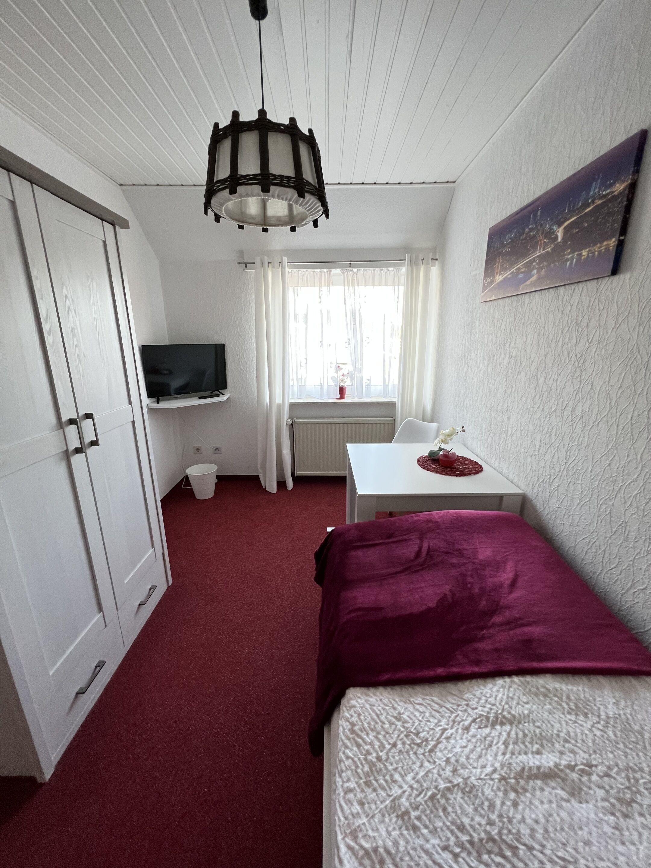Una habitación con cama, escritorio con silla, televisor, ventana con cortinas y un cuadro en la pared.