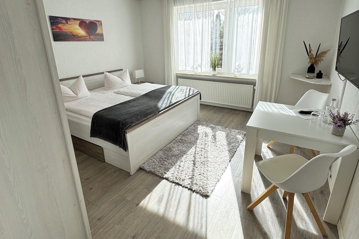 A bedroom with a bed, bedside table, desk, chair, and a window with curtains.