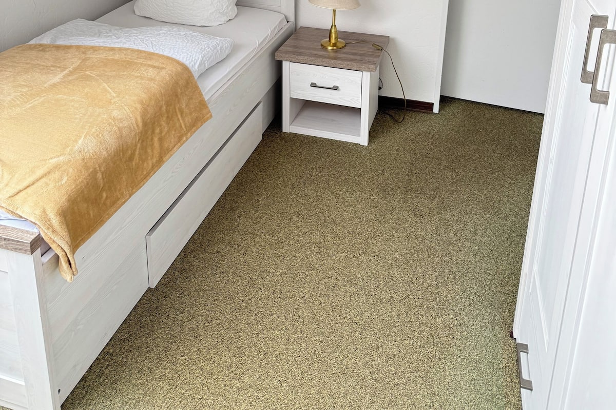 A small, white, single-bed room with a sloped ceiling, a bedside table with a lamp, and a door.