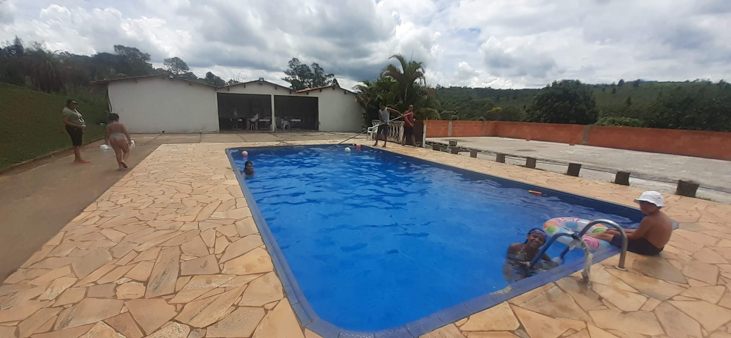A swimming pool with stone tiles, a covered area with tables, and a view of greenery in the background.