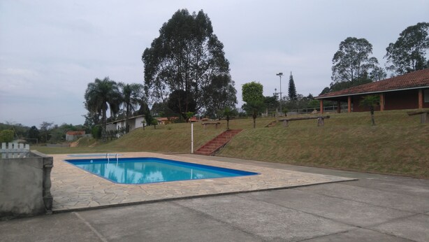 A swimming pool with a stone deck, surrounded by a grassy area and trees. - CHÁCARA LETÍCIA Season-Leisure and Events (Araçariguama)