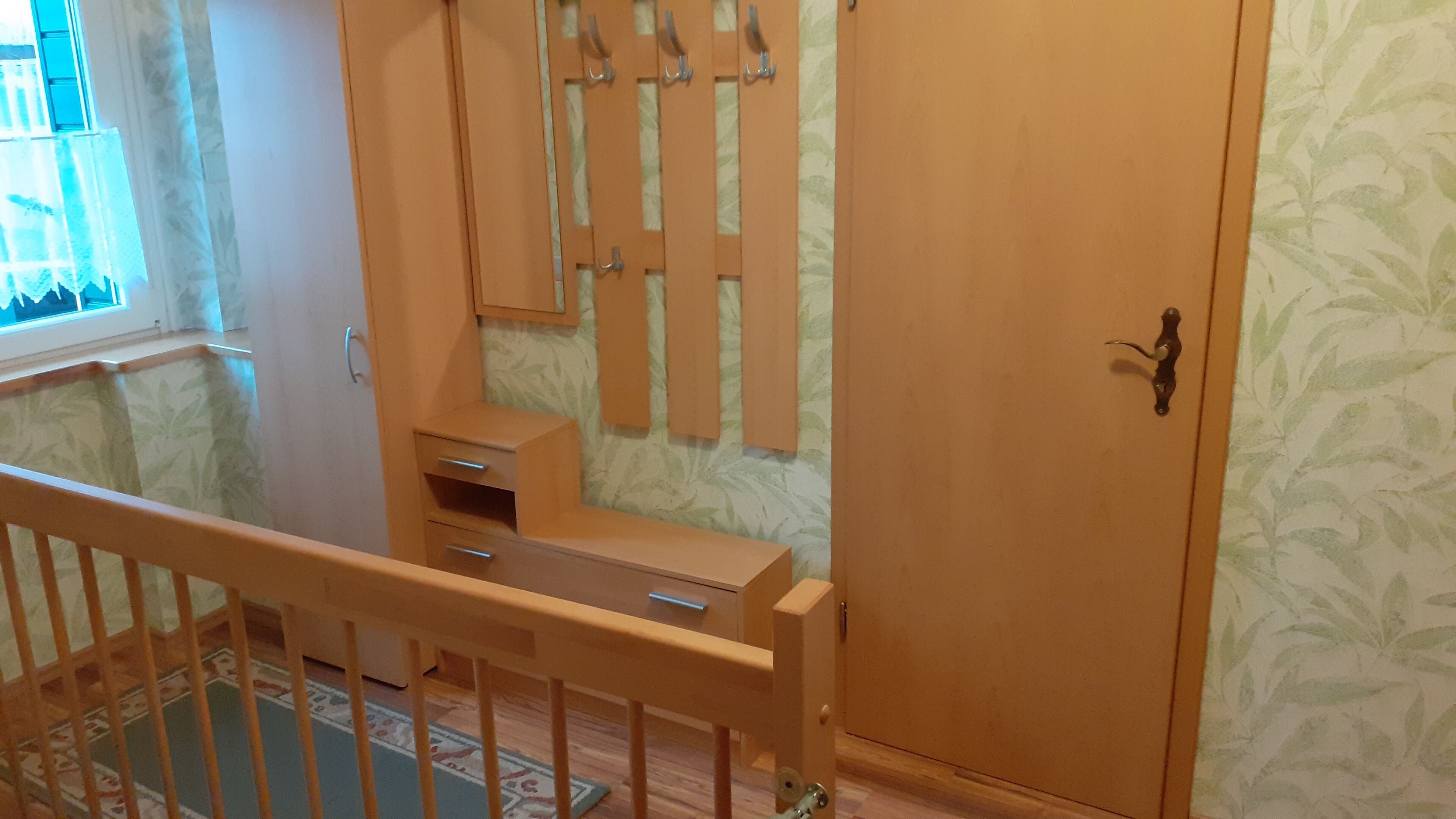 Ein Zimmer mit einem Holzschrank, einem kleinen Schreibtisch mit Schubladen, einem Fenster mit Vorhängen und einer Wand mit Musterung.