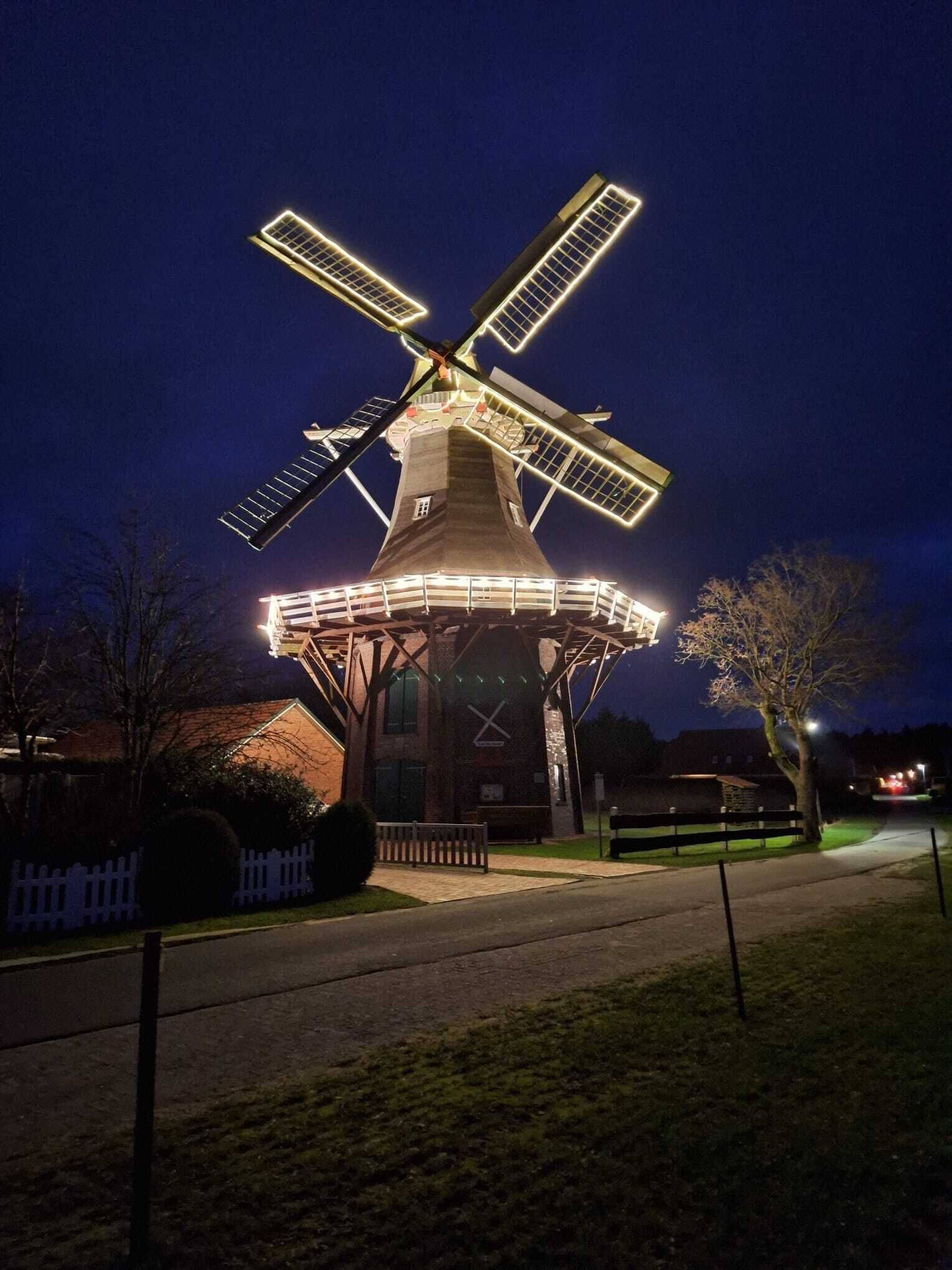 Un molino con aspas iluminadas y una base cubierta, rodeado por una valla y árboles.