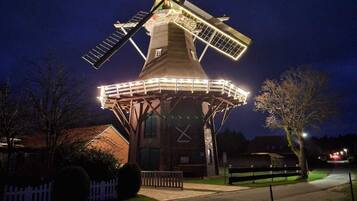 Un molino con aspas iluminadas y una base cubierta, rodeado por una valla y árboles.