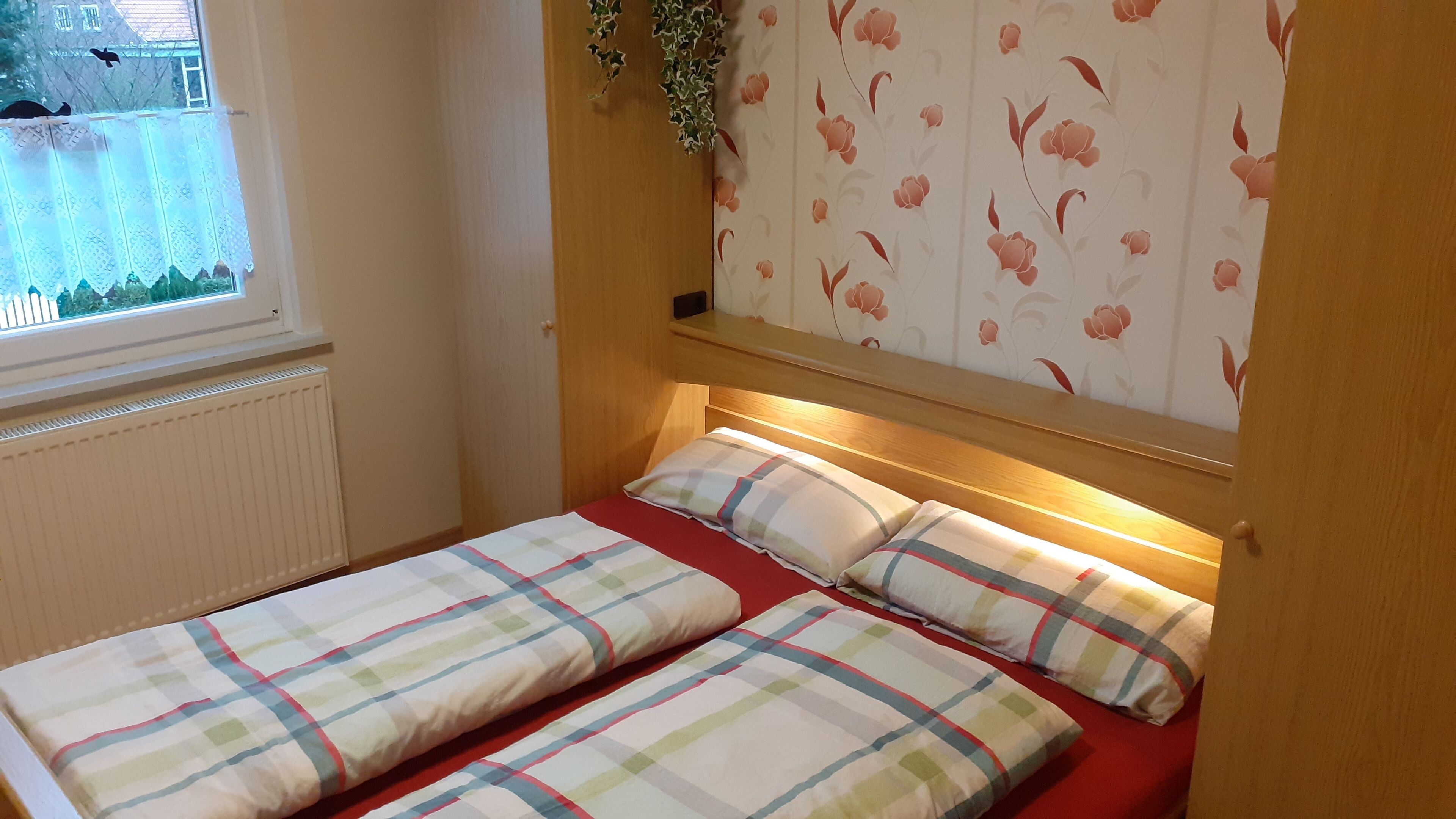 A small, cozy bedroom with a single bed, floral wallpaper, and a window with a view.