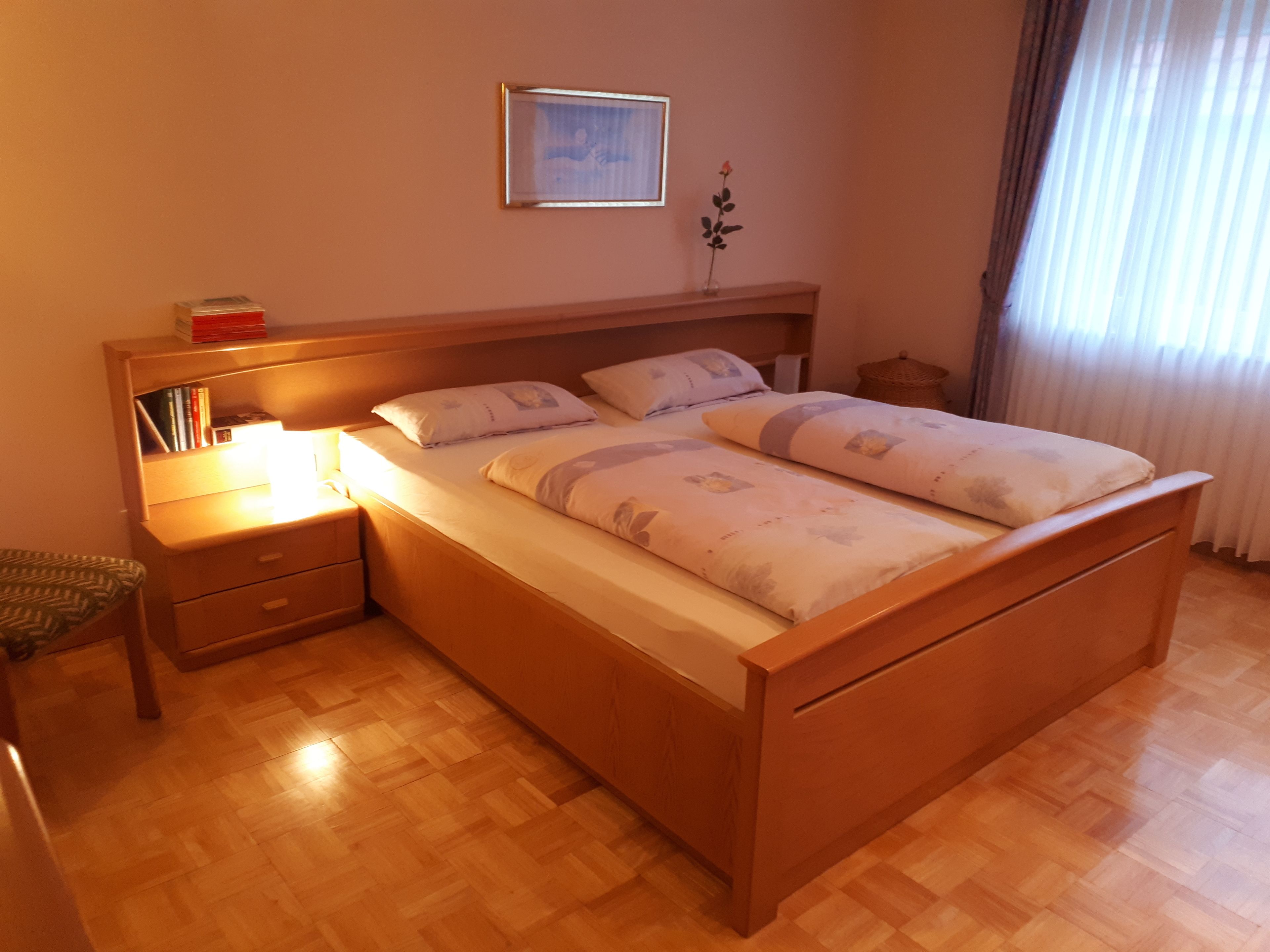 Ein hölzernes Bett mit Kopfteil, ein Nachttisch mit Lampe sowie ein kleinerer Nachttisch mit Büchern.