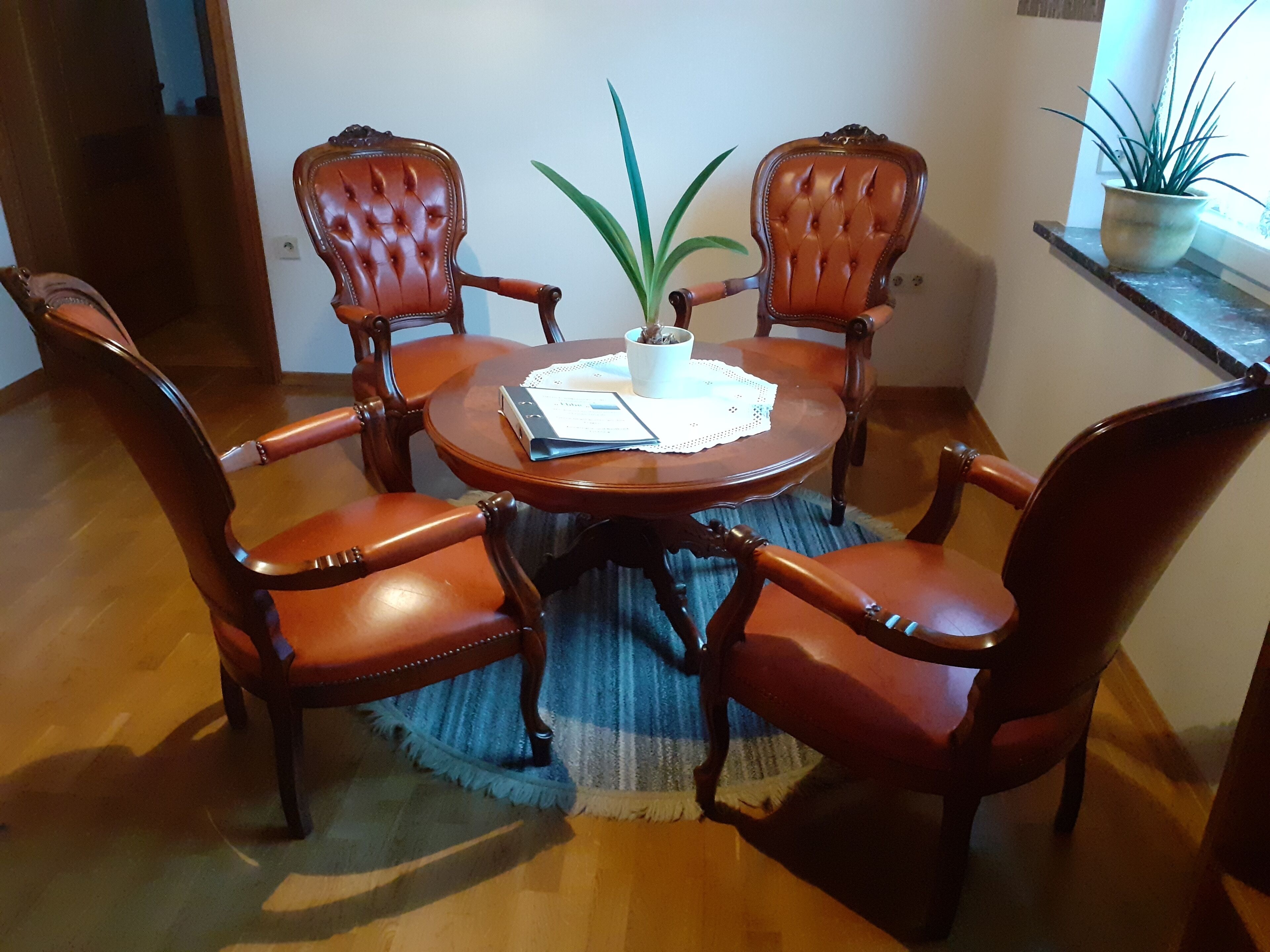 Een eetgedeelte met een ronde houten tafel, omringd door vier gestoffeerde stoelen. Op de tafel staat een potplant, en onder de tafel ligt een vloerkleed met blauw-witte patronen.