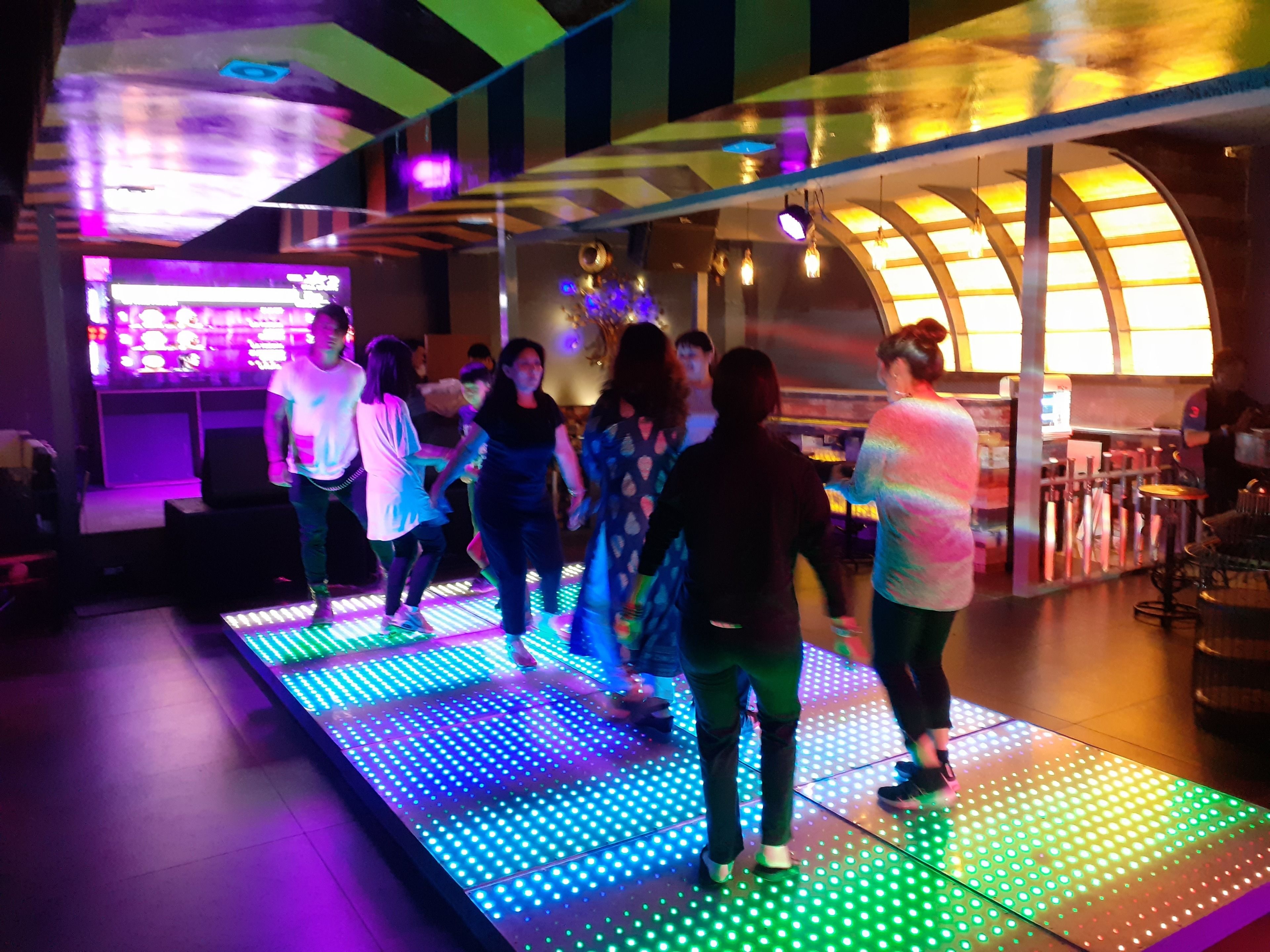 A group of people dancing on a brightly lit floor with a DJ booth in the background.