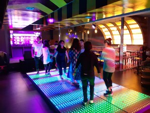 A group of people dancing on a brightly lit floor with a DJ booth in the background.