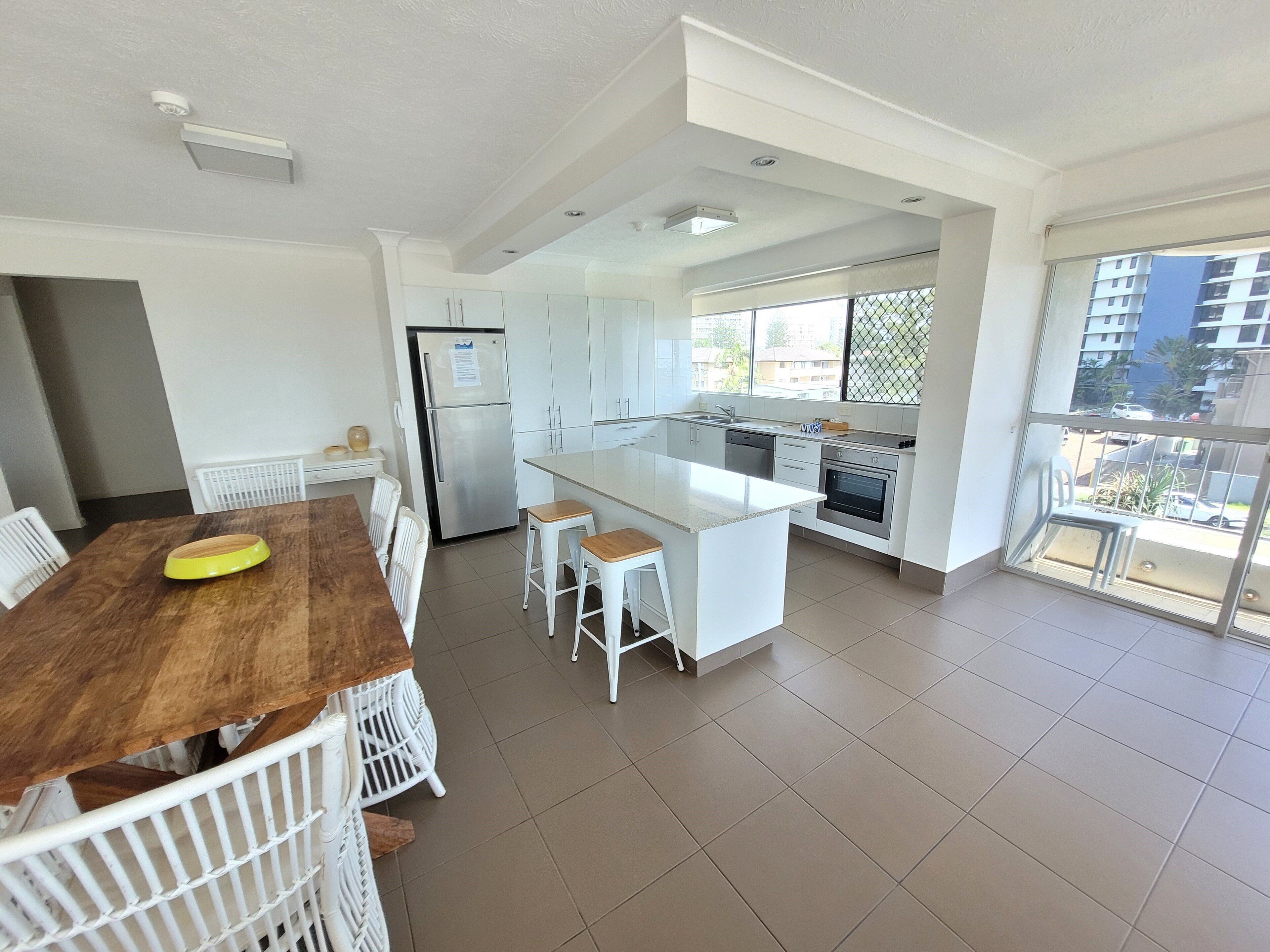 Une cuisine moderne avec des meubles blancs, un îlot central et un coin repas comprenant une table en bois et des chaises blanches.