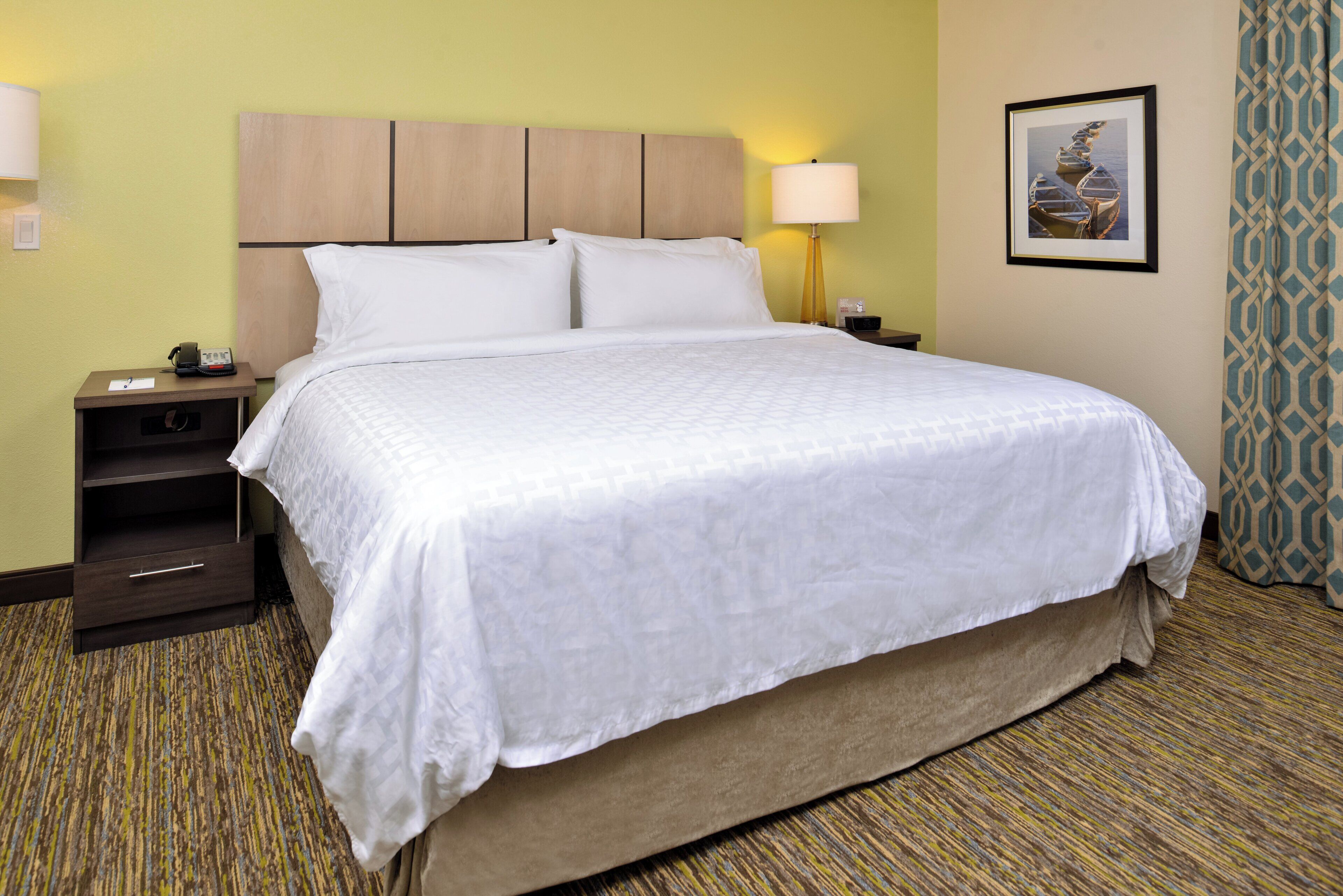 A hotel room with a bed, nightstand, lamp, and a framed picture on the wall.