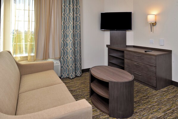 A hotel room with a sofa, a flat-screen TV mounted on a wall, a wooden cabinet with drawers, and a patterned curtain. - Candlewood Suites Lodi by IHG (Lodi)