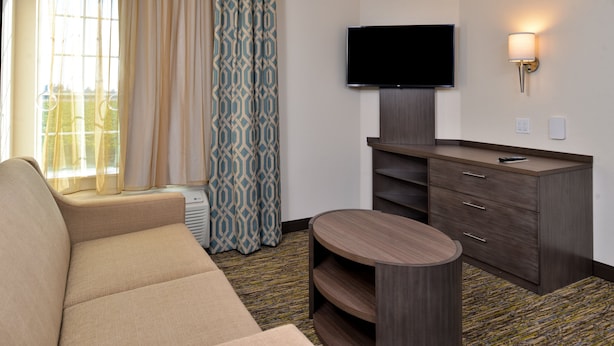 A hotel room with a beige sofa, a TV mounted on the wall, a wooden cabinet, and a patterned curtain.