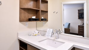 A hotel bathroom with a white sink, a mirror, a wooden shelf with toiletries, and a towel rack.